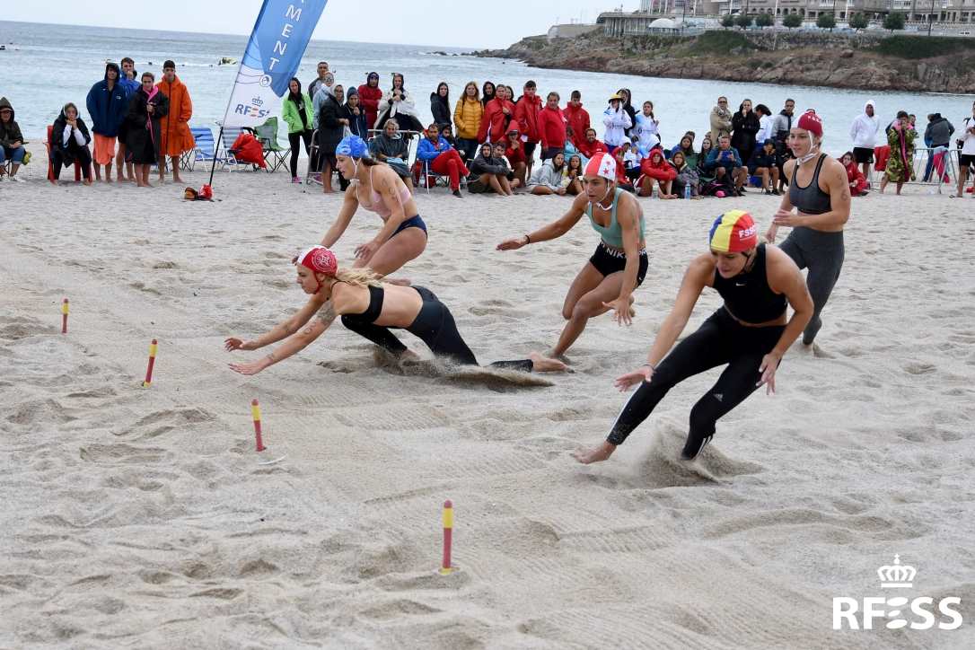 Varias socorristas se disputan los testigos en una serie de la prueba de banderas en la playa de Orzán, en A Coruña, durante la edición de la pasada temporada. AUTOR: Javier Sánchez-RFESS